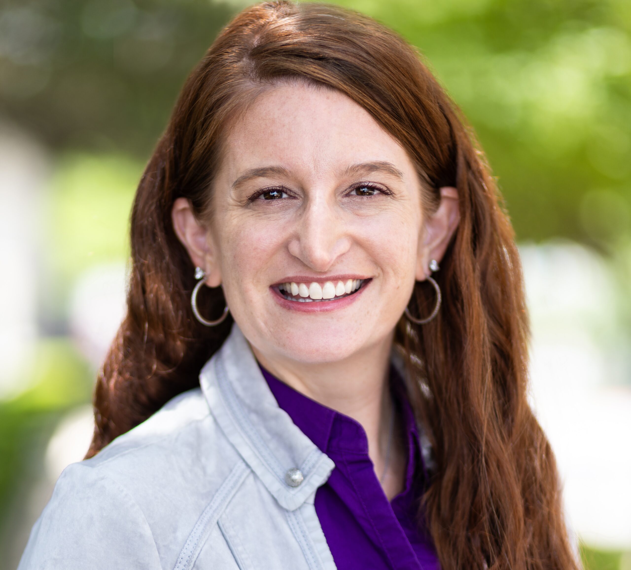 Woman smiling wearing a purple shirt and a light blue jacket with a green outdoor background