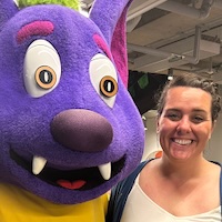 Woman smiling wearing a white shirt and black backpack next to the river bat mascot at ACC