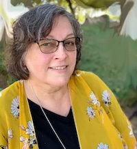 A woman smiling wearing a black shirt and yellow sweater with dark glasses and a green outdoor background