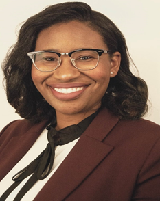 Image description: A Nigerian American woman smiling, wearing a maroon blazer, cream and black blouse, and black glasses.