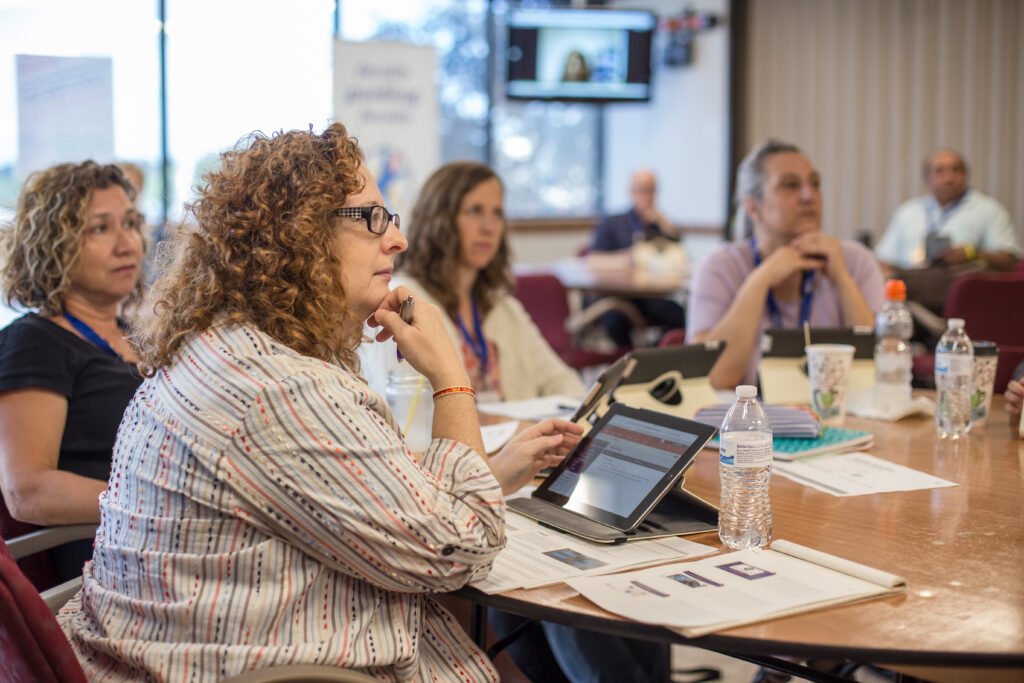 A diverse group of four individuals sitting at a table collaborating with a laptop, notebooks, and printed documents in a brightly lit educational setting