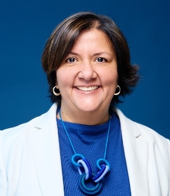 Headshot of Dr. Karina Hernandez smiling, with short dark hair, wearing a white blazer and a blue necklace against a blue background.